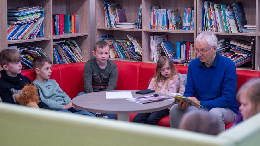 Ein älterer Mann mit Brille und blauem Pullover sitzt in einer Bibliotheksecke auf einem roten Sofa und liest vier Grundschulkindern aus einem Buch vor. Die Kinder hören aufmerksam zu. Im Hintergrund befinden sich helle Holzregale voller Kinderbücher.